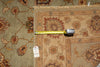 Load image into Gallery viewer, A close-up of a patterned area rug in beige, brown, and rust tones, with a yellow tape measure laid across the corner and a white inventory tag attached.