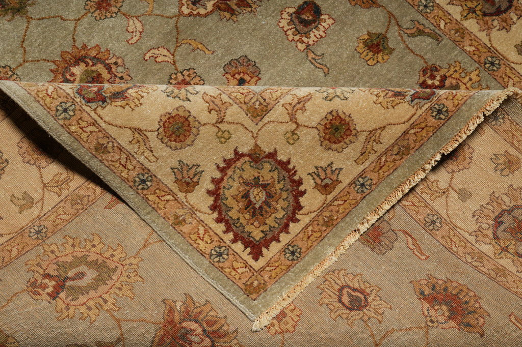 A close-up of a patterned area rug with a folded corner revealing the underside. The top surface shows a muted sage green field with floral motifs in beige, rust, and brown.