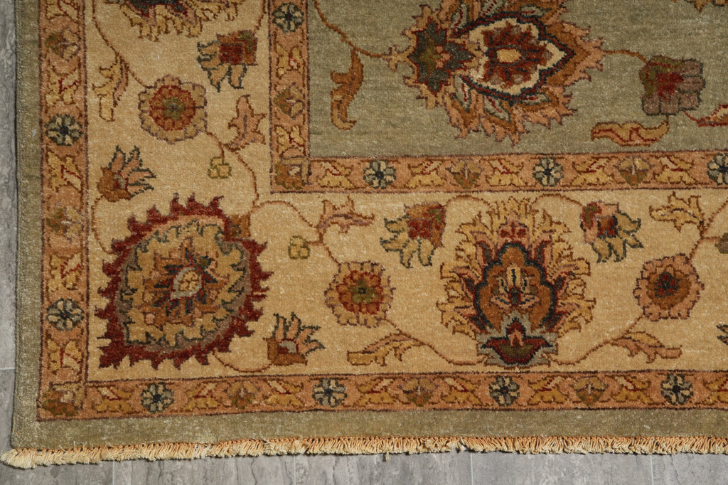 A close-up of a traditional patterned rug corner showing beige, olive green, rust red, and brown floral motifs with a fringed edge on a gray floor.