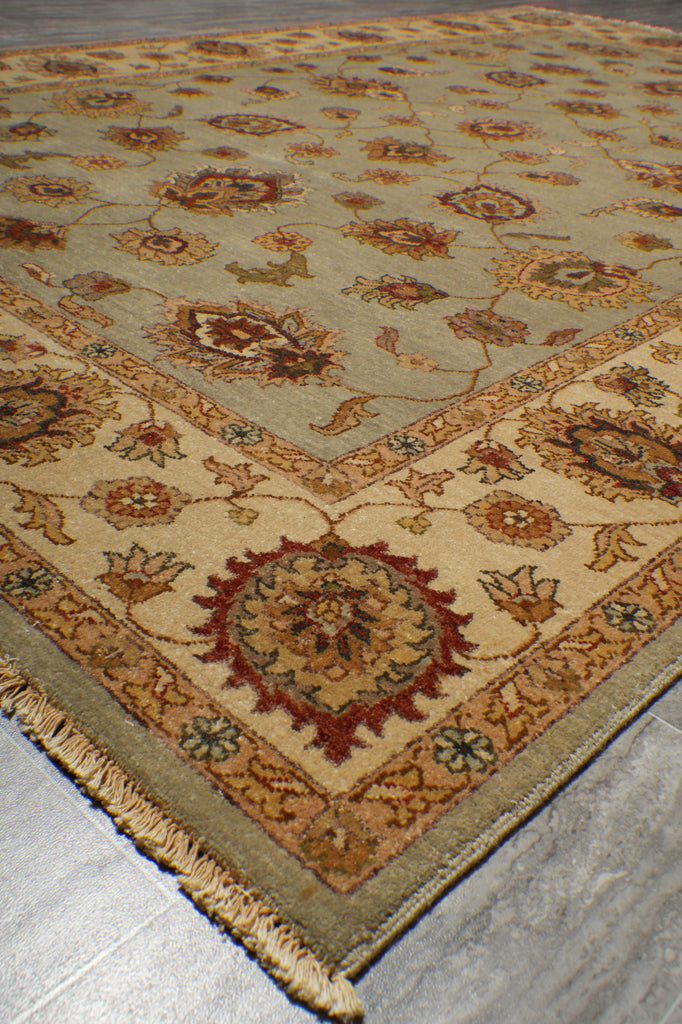 A traditional oriental-style area rug with a beige and olive green base, featuring intricate floral motifs and a detailed border, laid out on a gray tiled floor.