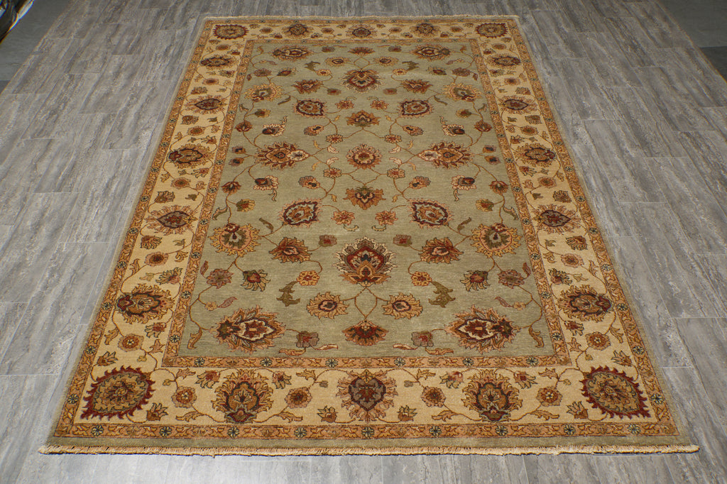 A rectangular area rug with an ornate floral and medallion pattern, laid out on a gray wood-look floor. The rug features a sage green center field, a beige inner border, and a darker brown outer border.