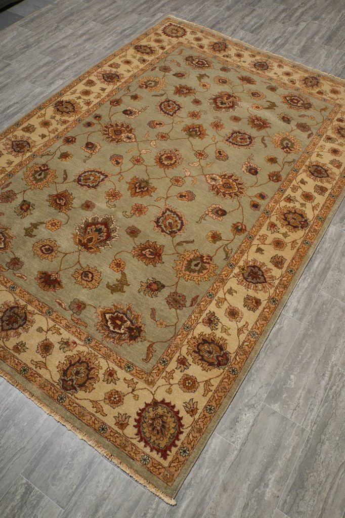 A rectangular area rug with an intricate floral and medallion pattern, shown from an elevated angle on a gray wood-look floor. The rug features a light sage green center field, a beige inner border, and a warm rust-orange outer border.