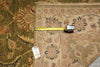 Load image into Gallery viewer, A beige patterned area rug with floral motifs is shown with a yellow tape measure laid across it for scale, alongside another darker green patterned rug. A small inventory tag is attached near the corner.