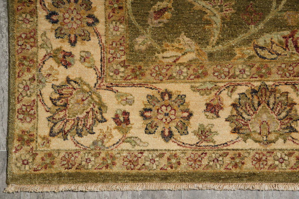 Close-up of an ornate area rug corner showing a cream field with floral motifs and a muted olive-green border, photographed on a gray floor.