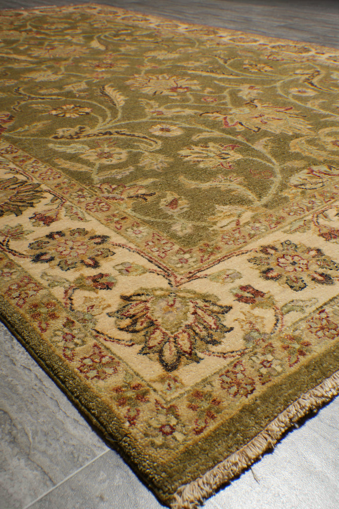 A close-up view of a traditional patterned area rug with a beige and olive green base, featuring floral motifs and fringed edges, laid out on a gray tiled floor.
