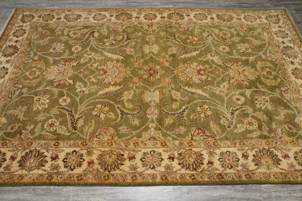 An ornate traditional area rug with a floral and vine pattern in olive green, beige, and rust red tones, laid out on a gray floor.