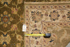 Load image into Gallery viewer, A close-up of a patterned area rug corner with a tape measure placed along the edge to show dimensions. The rug has a beige and brown base with floral motifs in muted blue, green, and rust tones.