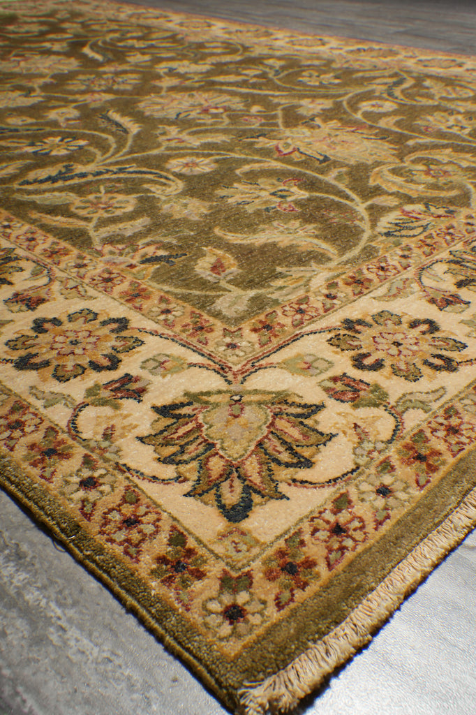 A close-up view of an ornate area rug corner, showing a traditional floral and vine pattern in beige, olive green, brown, and muted red tones with a fringed edge on a gray floor.