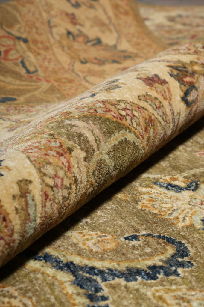 A close-up of a rolled section of an ornate area rug showing its detailed vintage-style pattern in beige, gold, brown, and navy blue.