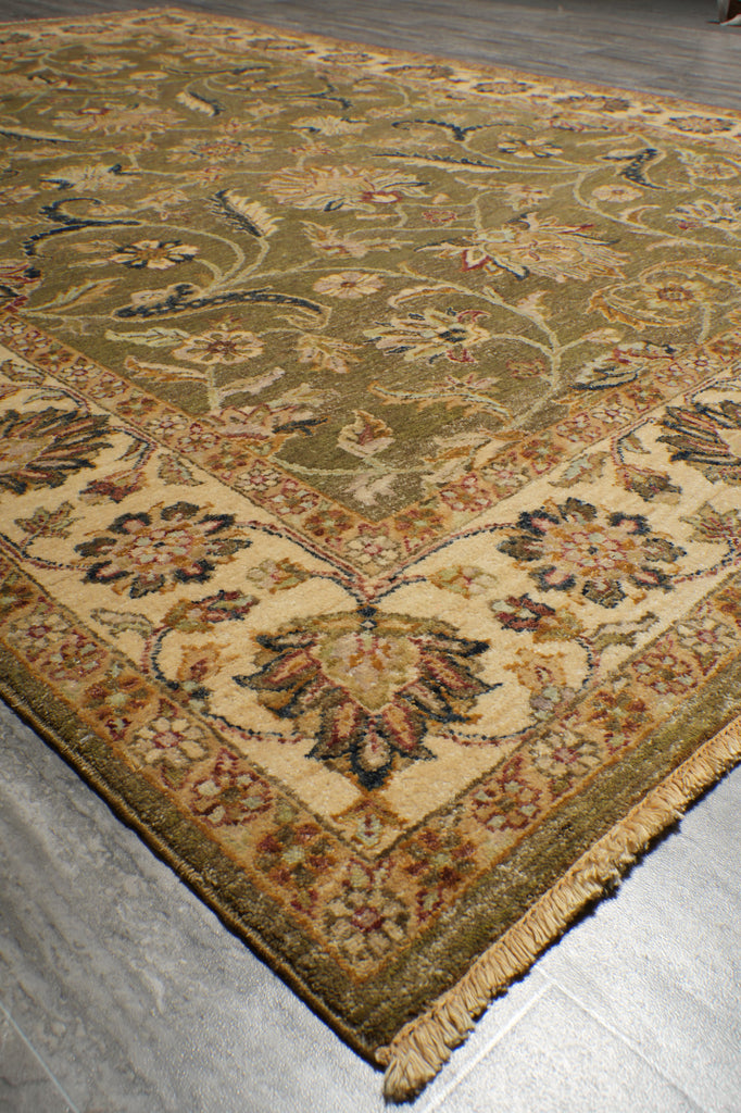 An ornate traditional area rug with a beige and olive-green base, featuring intricate floral motifs and a detailed border, shown on a gray wood-look floor.