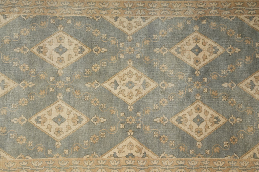 A close-up of a patterned rug with a muted blue-gray field, beige diamond medallions, and tan floral motifs, plus a decorative border along the top and bottom.