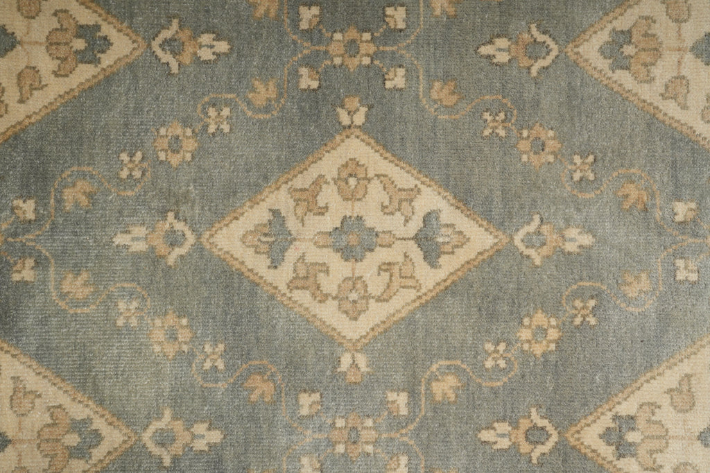A close-up of a traditional patterned rug featuring a dusty slate blue-gray background with beige and muted gold geometric diamond motifs and floral details.