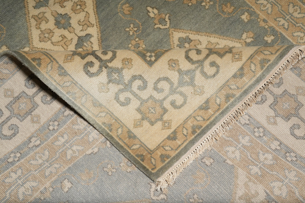 A close-up of a folded area rug showing its traditional pattern and fringed edge. The rug features a muted sage green, beige, and light blue-gray color palette with intricate floral and scroll motifs.