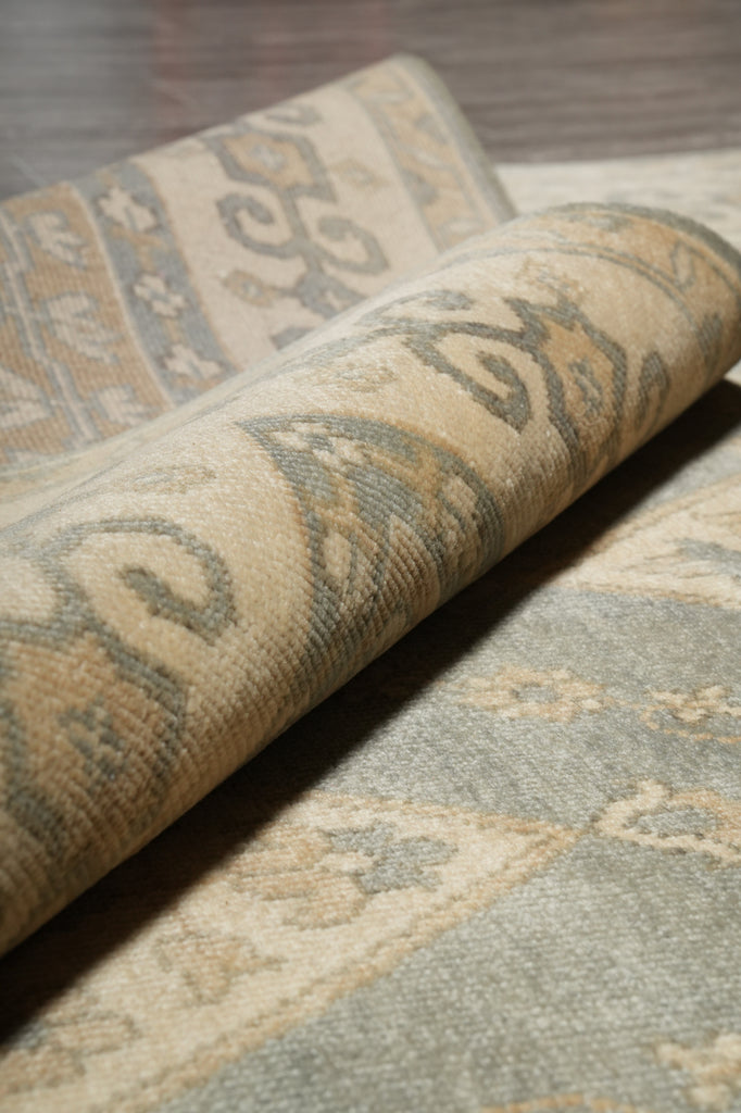 Close-up of a rolled area rug showing a traditional pattern in beige, gray, and muted blue-green tones.