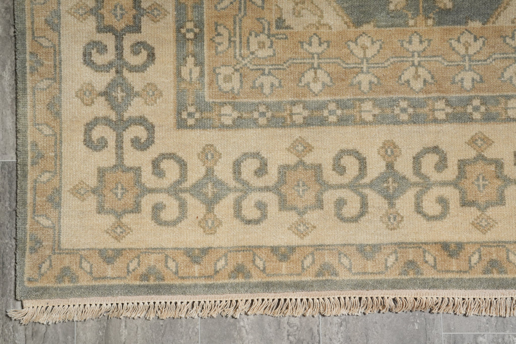 Close-up of a traditional patterned rug corner showing beige, gray, and muted blue-gray motifs with fringe along the bottom edge.