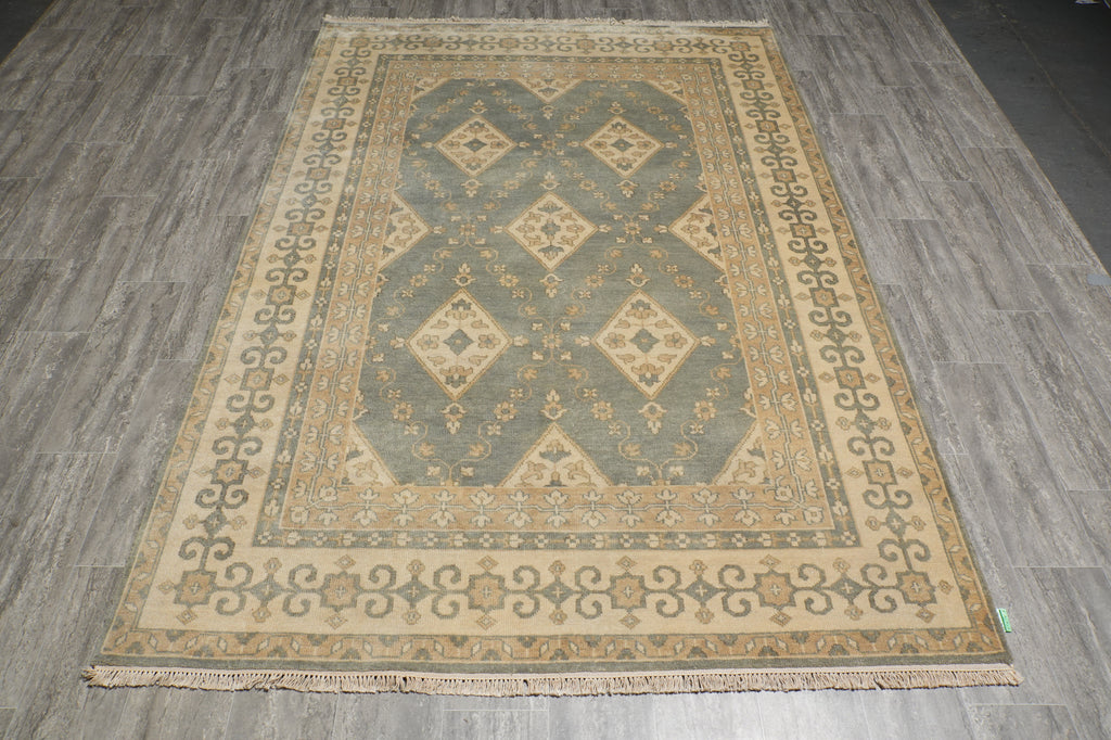 A rectangular area rug with a traditional geometric pattern in beige, gray, and muted green, laid out on a gray wood-look floor.