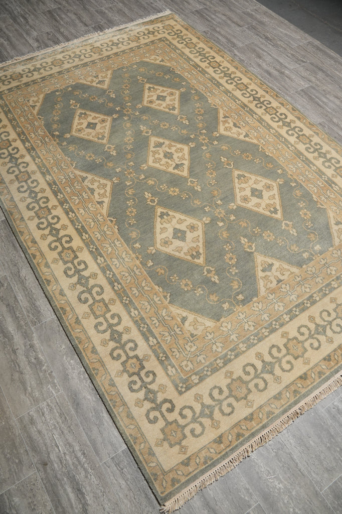A rectangular area rug with a traditional geometric pattern in beige, gray, and muted green, shown laid out on a gray wood-look floor.