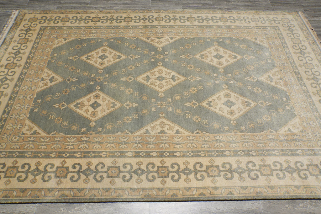 A rectangular area rug with a traditional geometric pattern in muted sage green, beige, and brown tones, laid out on a dark wood floor.