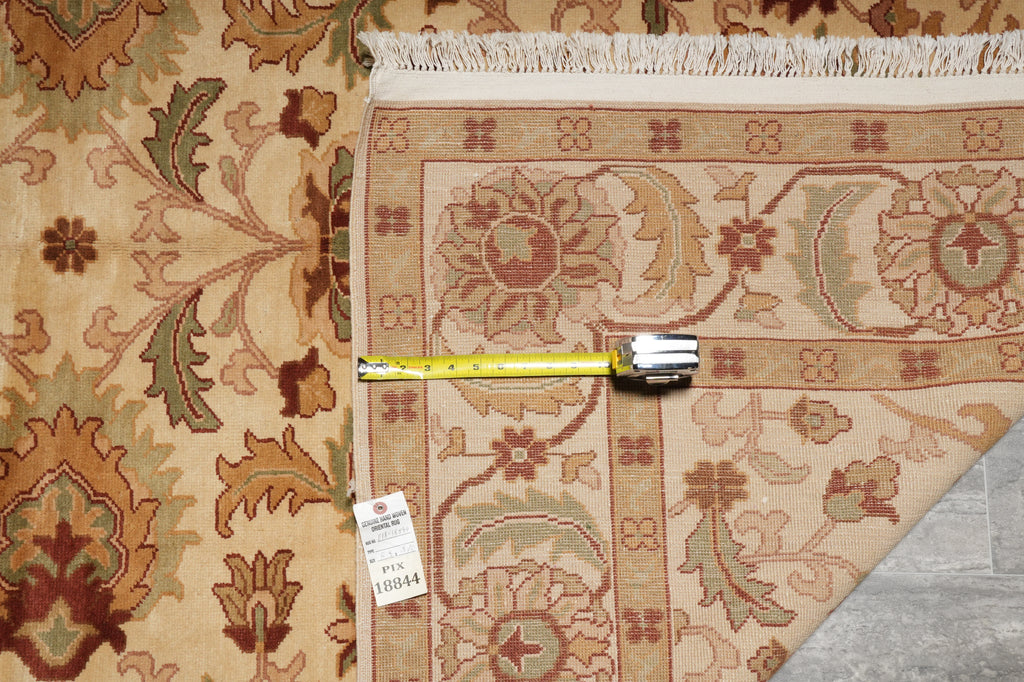 A close-up of a patterned area rug with a beige base and floral motifs in brown, rust, and muted green, shown with a yellow tape measure and price tag on a gray floor.