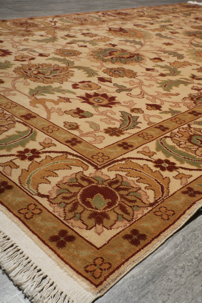 A close-up of a traditional patterned area rug with cream, tan, brown, and muted green floral motifs and a fringed edge, shown against a gray background.