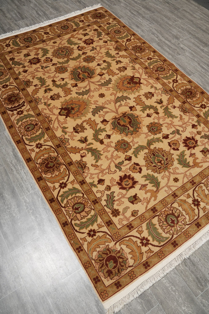 A rectangular traditional area rug with an intricate floral and medallion pattern, laid out on a gray wood-look floor. The rug features a warm beige center, a brown border, and accents of green, rust, and cream in the detailed motifs and fringe.