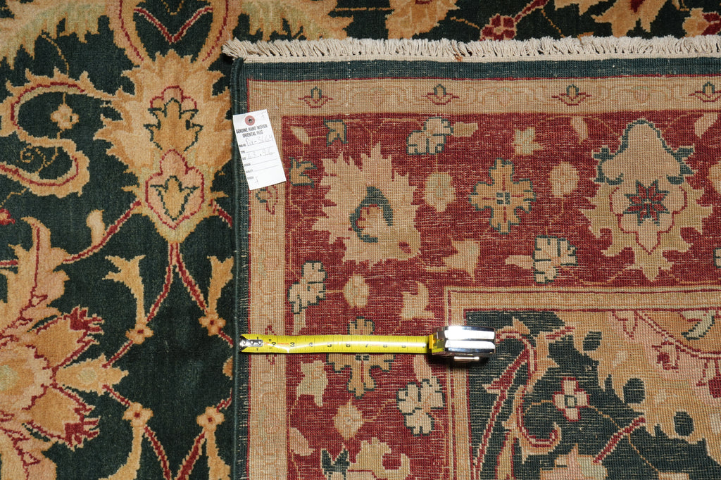 A folded area rug with a traditional floral pattern, showing a red field, beige borders, and a dark green outer section. A yellow tape measure lies horizontally across the fold to indicate size, and a white product tag is attached near the top left corner.