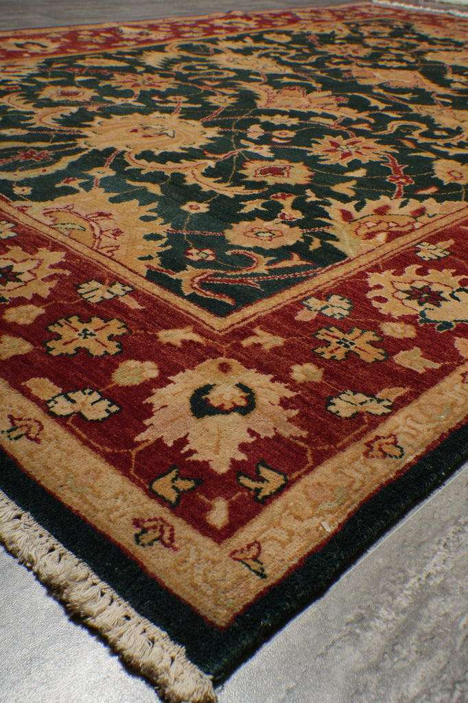 A close-up of a traditional patterned area rug showing its corner detail, featuring a dark green field, a deep red border, and cream floral motifs.