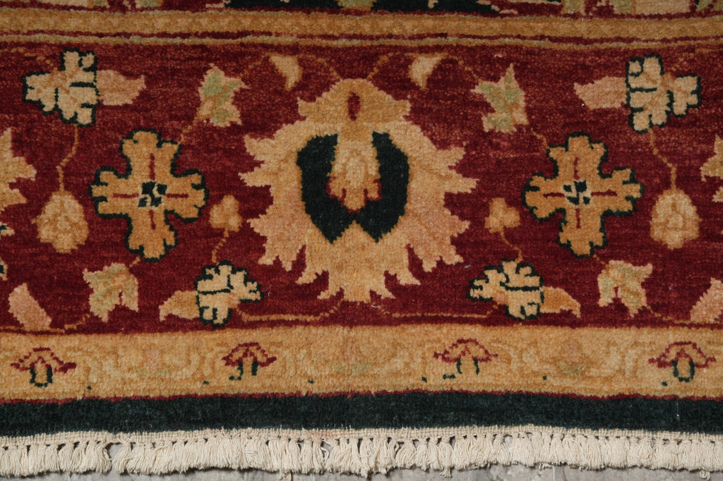 Close-up of a patterned area rug with a deep red field, beige floral motifs, a central dark green medallion, and cream fringe along the bottom edge.
