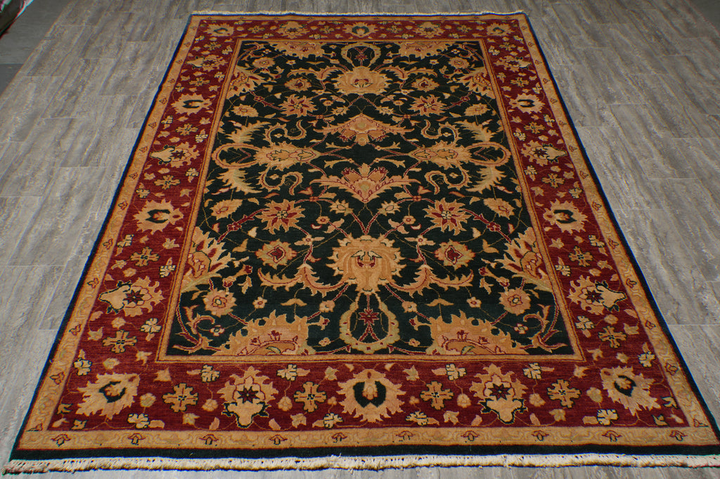 A traditional oriental area rug with an intricate floral pattern, featuring a dark green center, red borders, and beige accents, laid out on a gray wood-look floor.