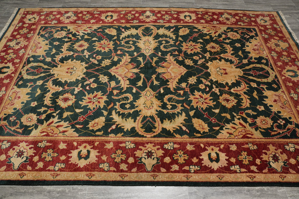 A rectangular area rug with an ornate floral and vine pattern, featuring a dark green center field, red borders, and beige accents, laid out on a gray floor.