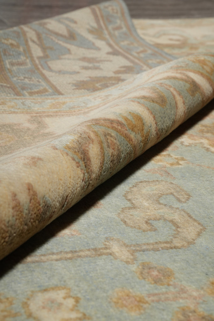 Close-up of a patterned area rug with a rolled corner showing the pile and border design in beige, cream, and muted blue tones.