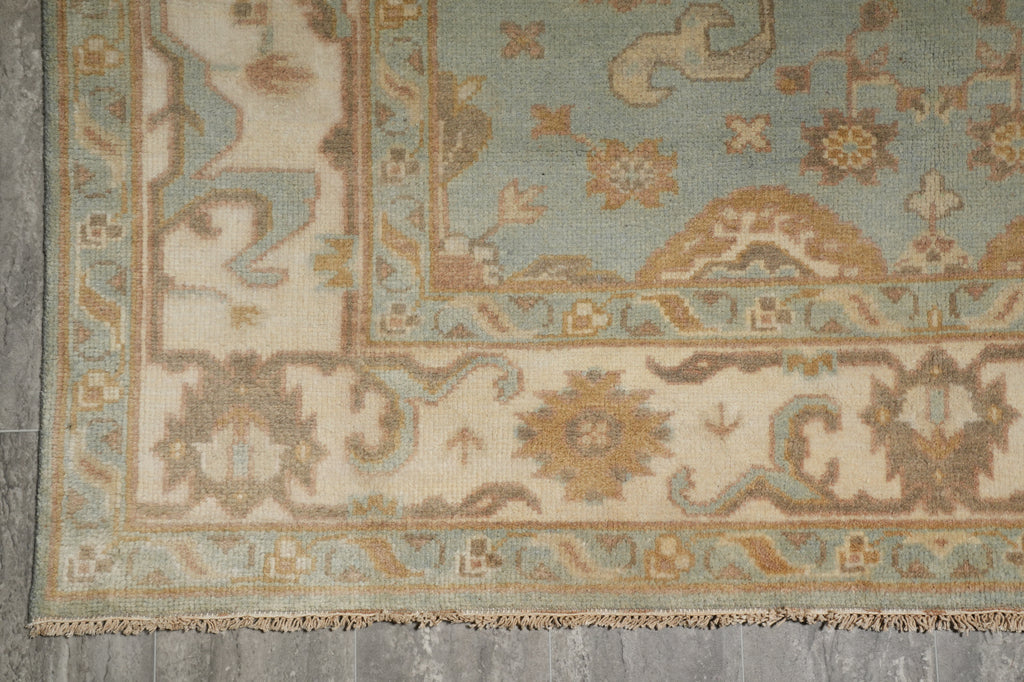A close-up of a traditional patterned rug corner, showing a dusty blue field, beige and cream borders, and fringed edges on a gray floor.