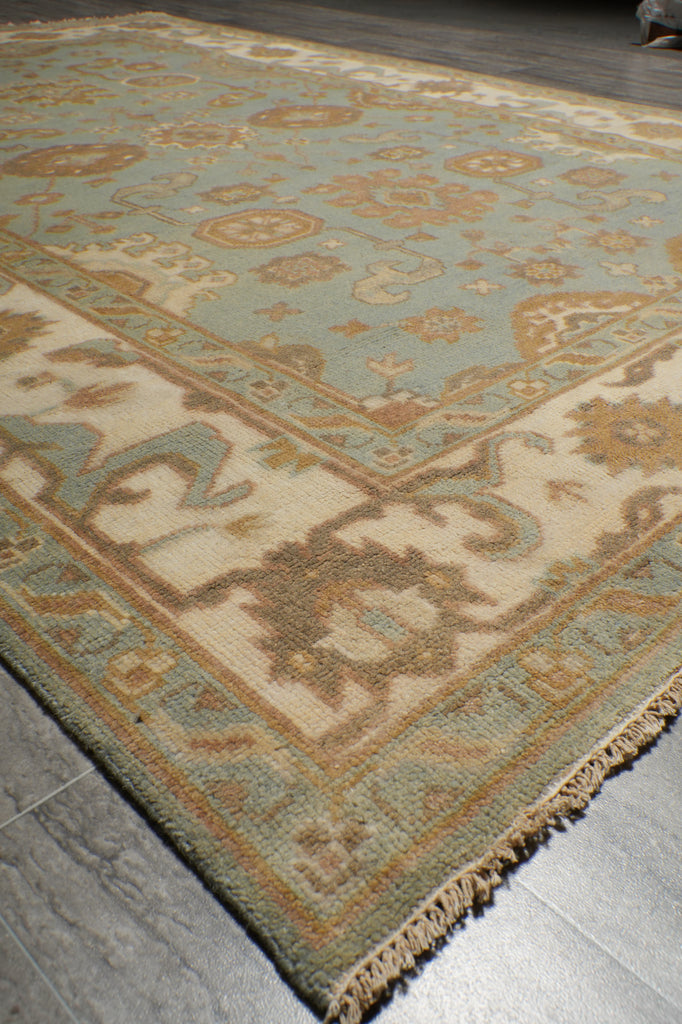 A close-up of a traditional patterned area rug with a light blue-green field, beige borders, and cream central motifs, shown on a gray tiled floor.