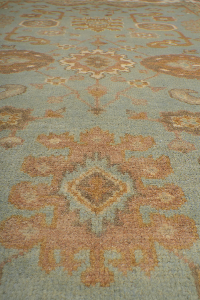A close-up of a patterned rug or textile with a muted sage green background and large peach, tan, and beige floral or medallion motifs.