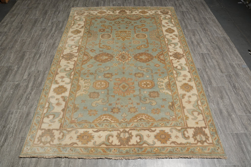 A rectangular traditional area rug with an ornate floral and medallion pattern, featuring a light blue center field and cream borders, laid out on a gray wood-look floor.