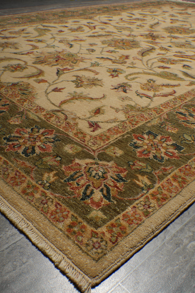 A traditional oriental-style area rug with a beige center field, green border, and multicolored floral motifs, shown laid out on a gray floor.