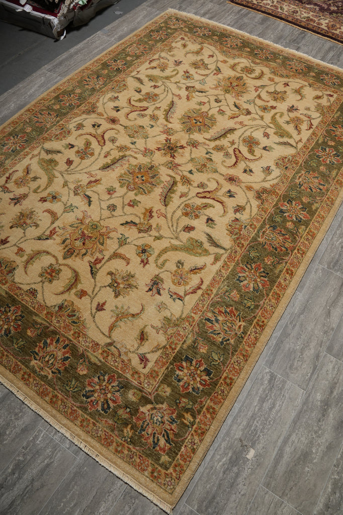 An ornate rectangular area rug with a beige center and detailed floral motifs, bordered in green, rust, and cream, shown on a gray tiled floor.