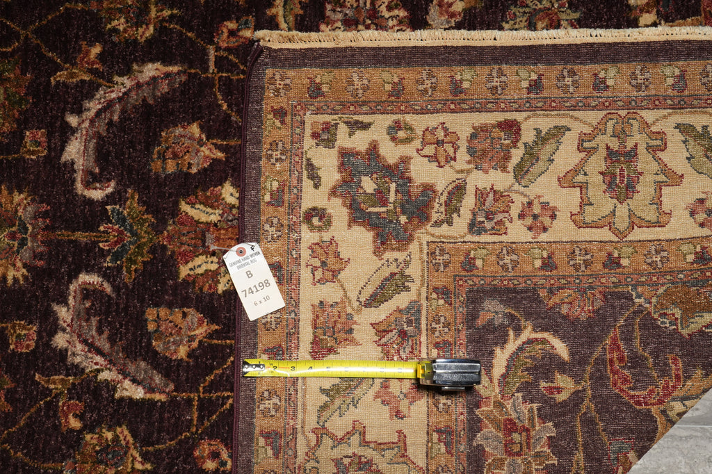 A close-up of a patterned area rug with a cream and brown floral design, shown alongside another dark rug. A yellow tape measure is laid across the border to indicate size, and a white tag is attached near the edge.