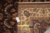 Load image into Gallery viewer, A close-up of a patterned area rug with a cream and brown floral design, shown alongside another dark rug. A yellow tape measure is laid across the border to indicate size, and a white tag is attached near the edge.