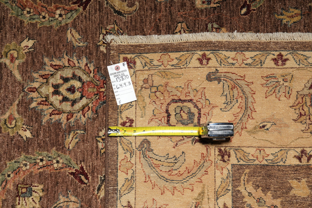 A close-up of an ornate patterned area rug with a beige central field, brown borders, and floral motifs in muted red, blue, and green tones. A yellow tape measure and a small white tag are placed on the rug to indicate scale and product details.