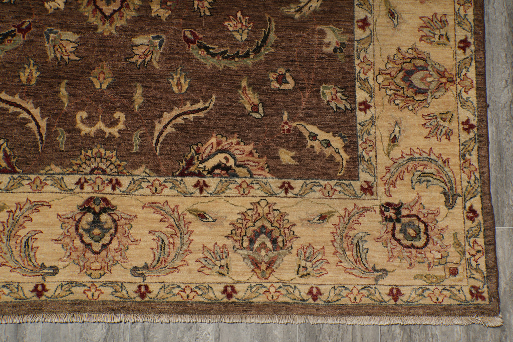 A close-up of a traditional patterned rug corner, showing a dark brown field with floral motifs and a beige border with ornate detailing, laid on a gray floor.