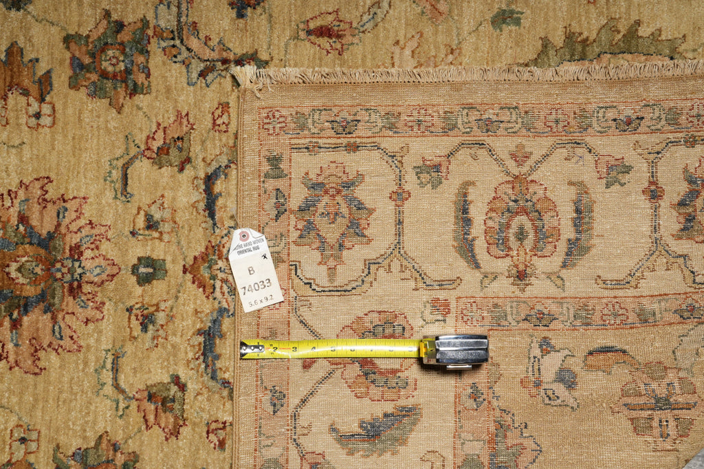 A close-up of a traditional patterned rug with a beige base and muted red, blue, and green floral motifs, shown with a yellow tape measure and a white tag on the floor.