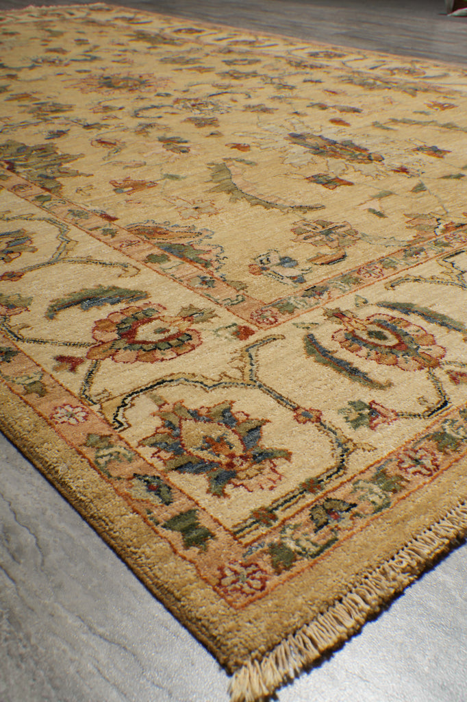 A close-up diagonal view of a traditional patterned area rug with a cream and beige base, featuring floral motifs in muted red, green, and blue, with a fringed edge.