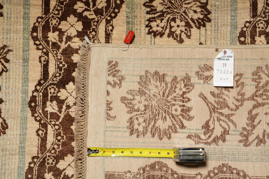 A close-up of a patterned area rug laid flat, showing a beige and brown floral design with fringe on one side. A yellow tape measure and a small white tag are placed on top of the rug for scale and identification.