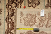 Load image into Gallery viewer, A close-up of a patterned area rug laid flat, showing a beige and brown floral design with fringe on one side. A yellow tape measure and a small white tag are placed on top of the rug for scale and identification.