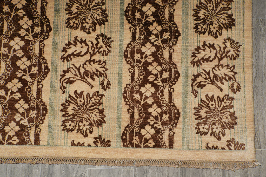A close-up of a patterned area rug with vertical stripes featuring dark brown floral and ornamental motifs on a beige background, with a fringed edge, laid on a gray floor.