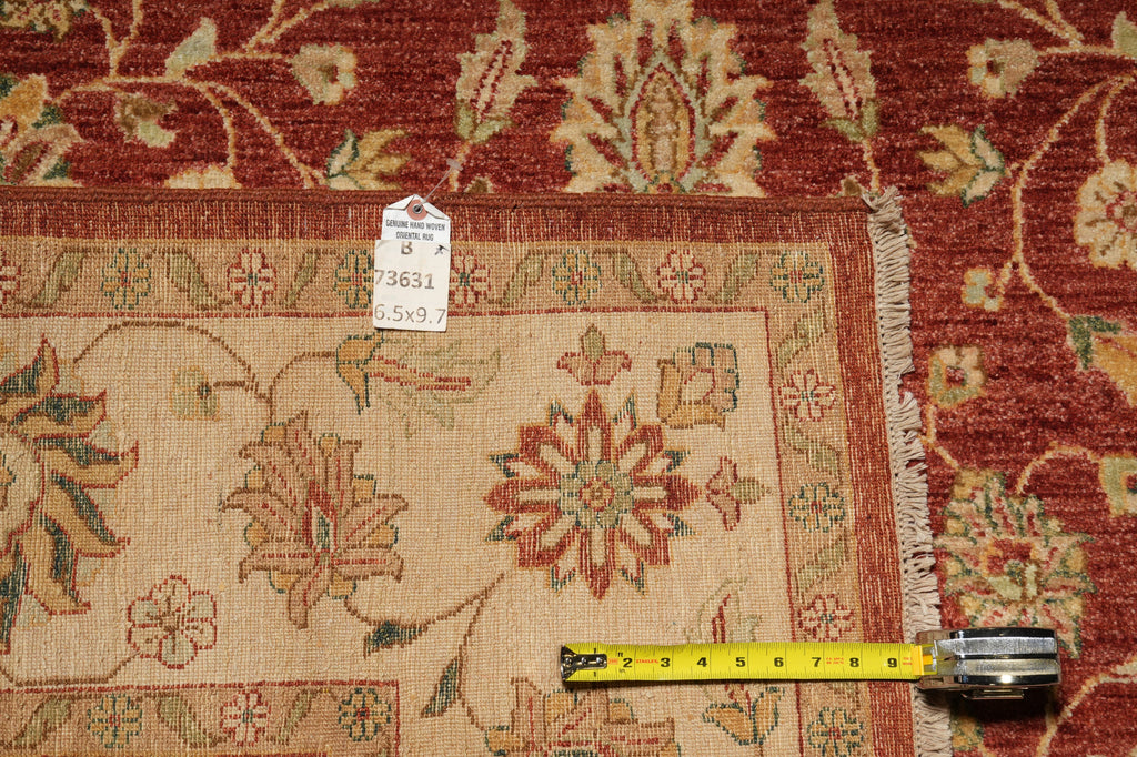 A close-up of a patterned area rug with a red outer field, beige inner field, and floral motifs. A yellow tape measure is placed along the fringe to show the rug's width, and a small product tag is attached near the center.
