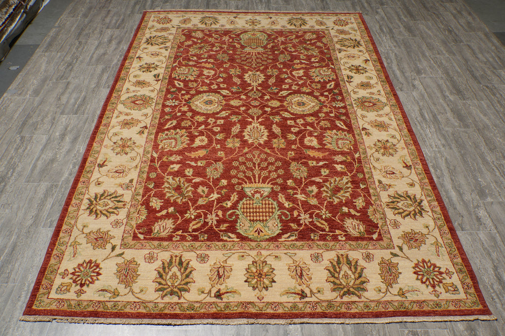 A rectangular traditional area rug with an ornate floral and botanical pattern, featuring a deep red central field, cream-colored borders, and accents of green, gold, and brown, displayed on a gray wood-look floor.