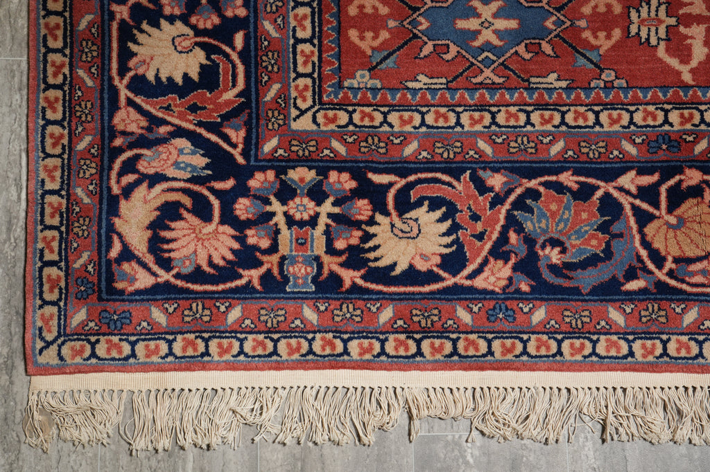 A close-up of an ornate patterned rug corner with cream fringe, featuring a dark navy field, red border, and floral motifs in beige, rust, and muted blue on a gray floor.