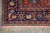 Load image into Gallery viewer, A close-up of an ornate patterned rug corner with cream fringe, featuring a dark navy field, red border, and floral motifs in beige, rust, and muted blue on a gray floor.
